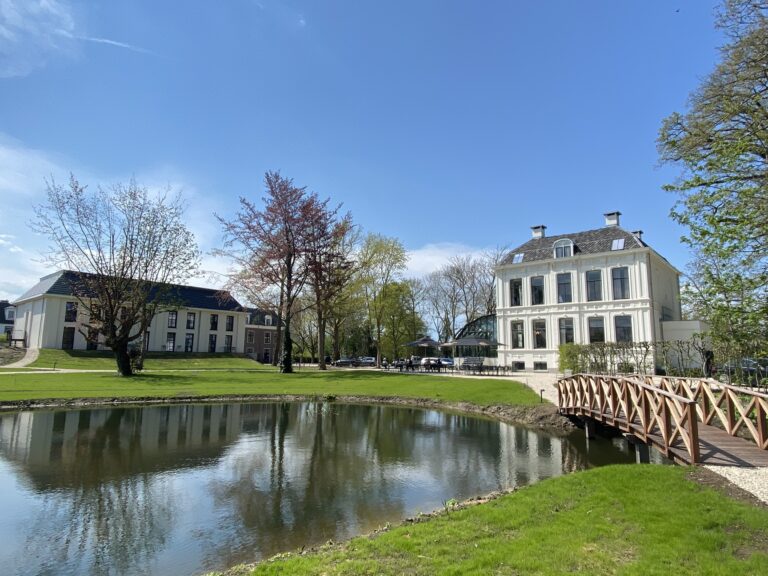 Stijlvol wit landhuis met terras, weerspiegeld in een vijver met houten brug op een zonnige dag.