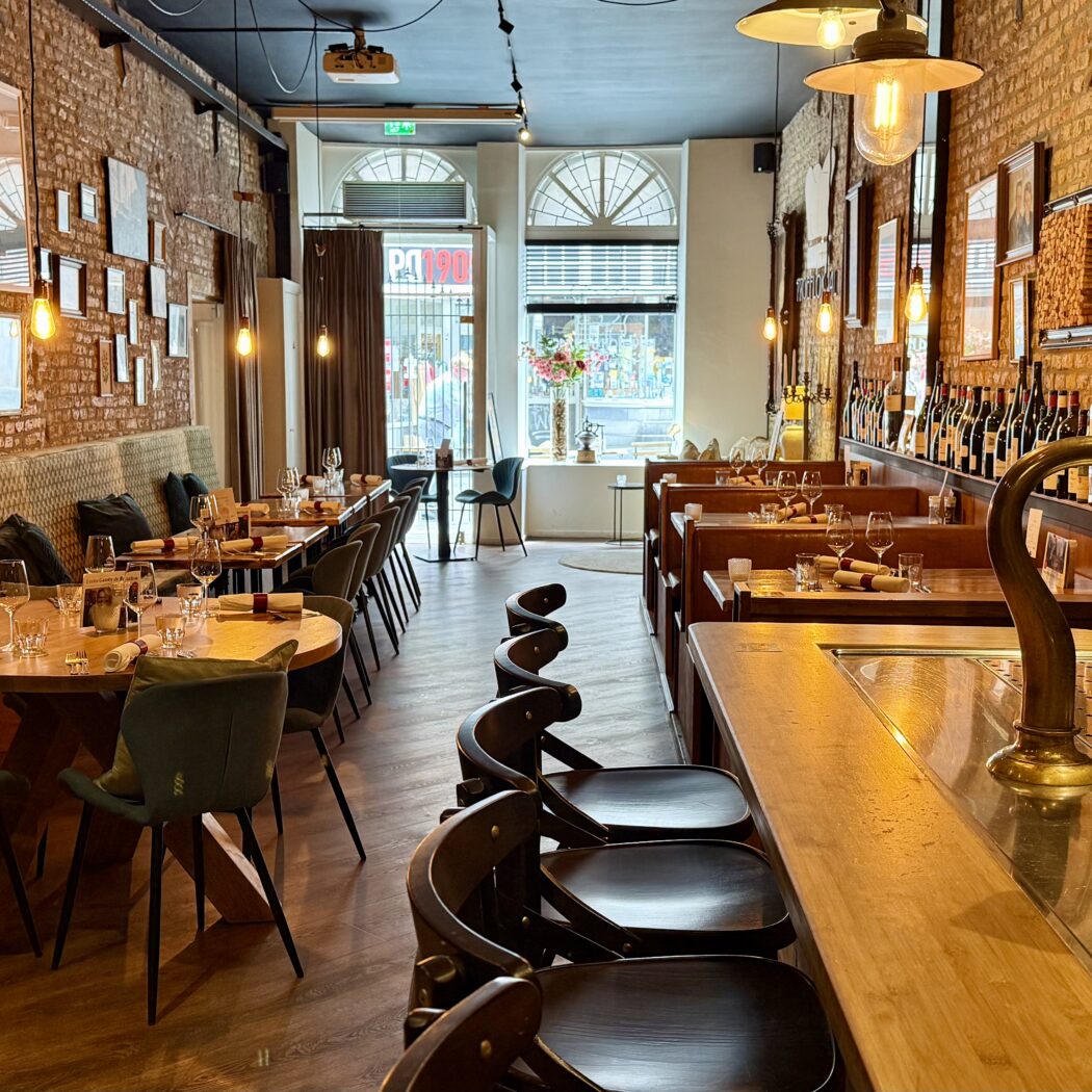 Restaurant interieur: houten tafels, stoelen en bar met biertap; bakstenen muur met wanddecoratie.