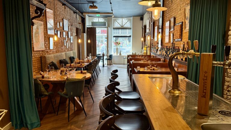 Restaurant interieur: houten tafels, stoelen en bar met biertap; bakstenen muur met wanddecoratie.
