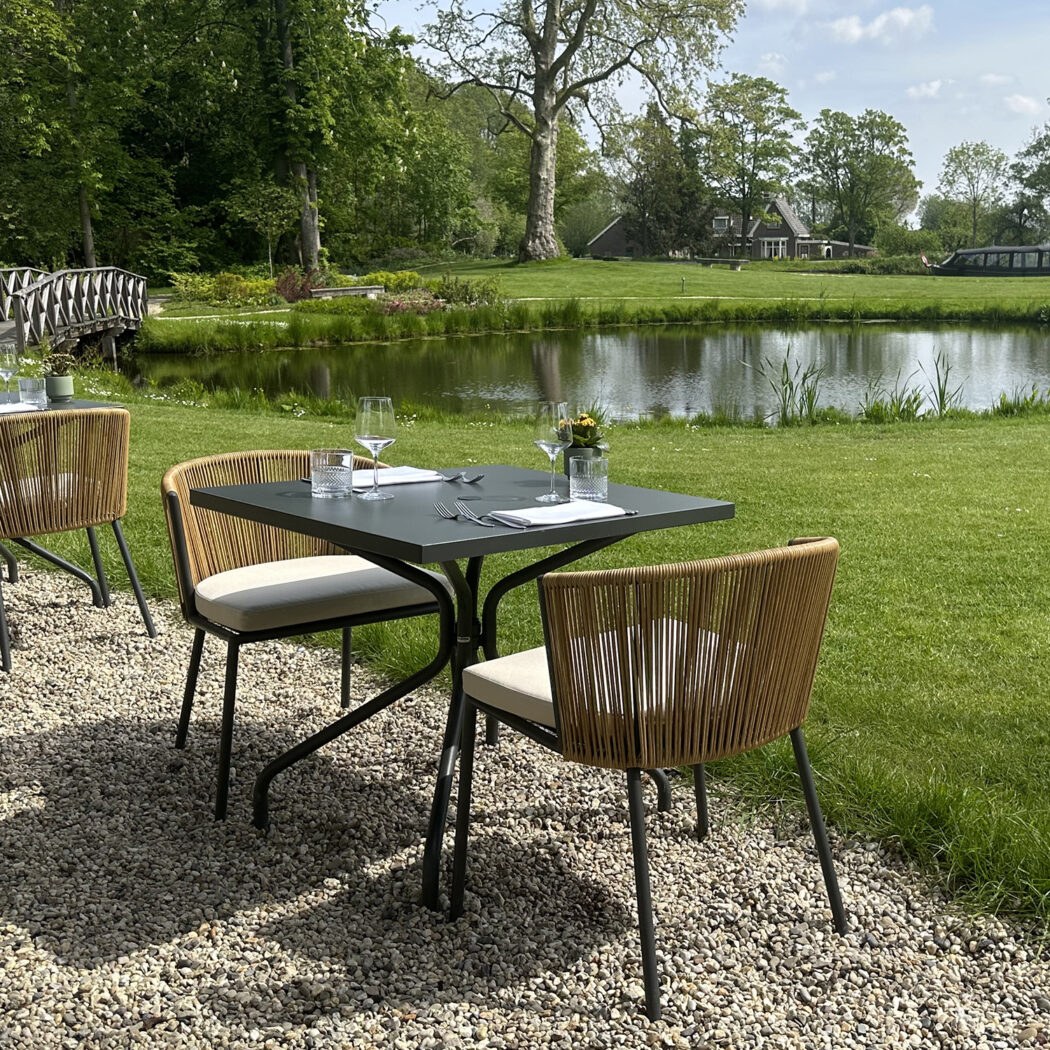 Gedekte buitentafels en moderne stoelen op een grindterras met uitzicht op een vijver, bomen en een brug.