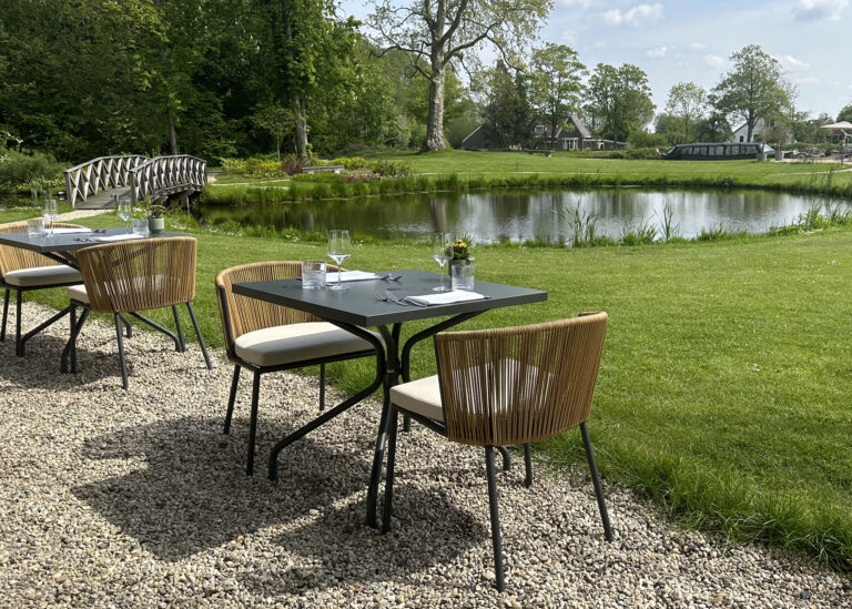 Gedekte buitentafels en moderne stoelen op een grindterras met uitzicht op een vijver, bomen en een brug.