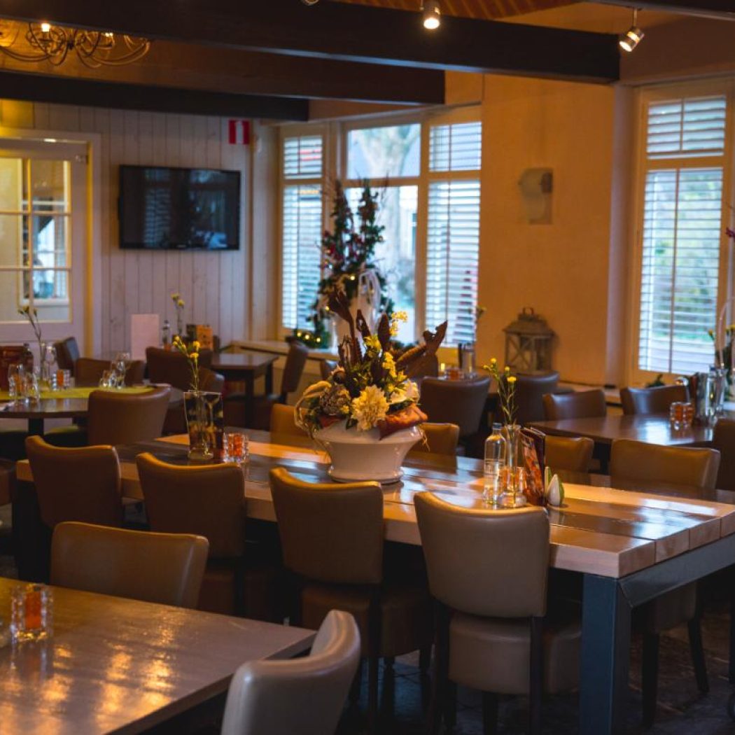 Sfeervol restaurantinterieur met lege houten tafels en bruine stoelen, versierd met kerstdecoraties en warme verlichting.