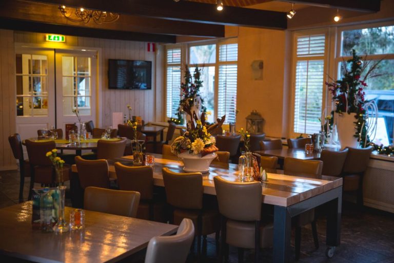 Sfeervol restaurantinterieur met lege houten tafels en bruine stoelen, versierd met kerstdecoraties en warme verlichting.