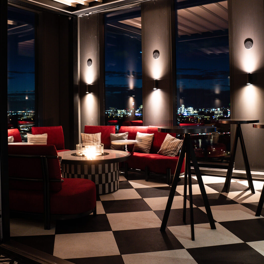 Rooftop lounge met rode zitmeubels, zwart-wit geblokte vloer, kaars op tafel en Delicious op het raam, met nachtelijk stadsuitzicht.