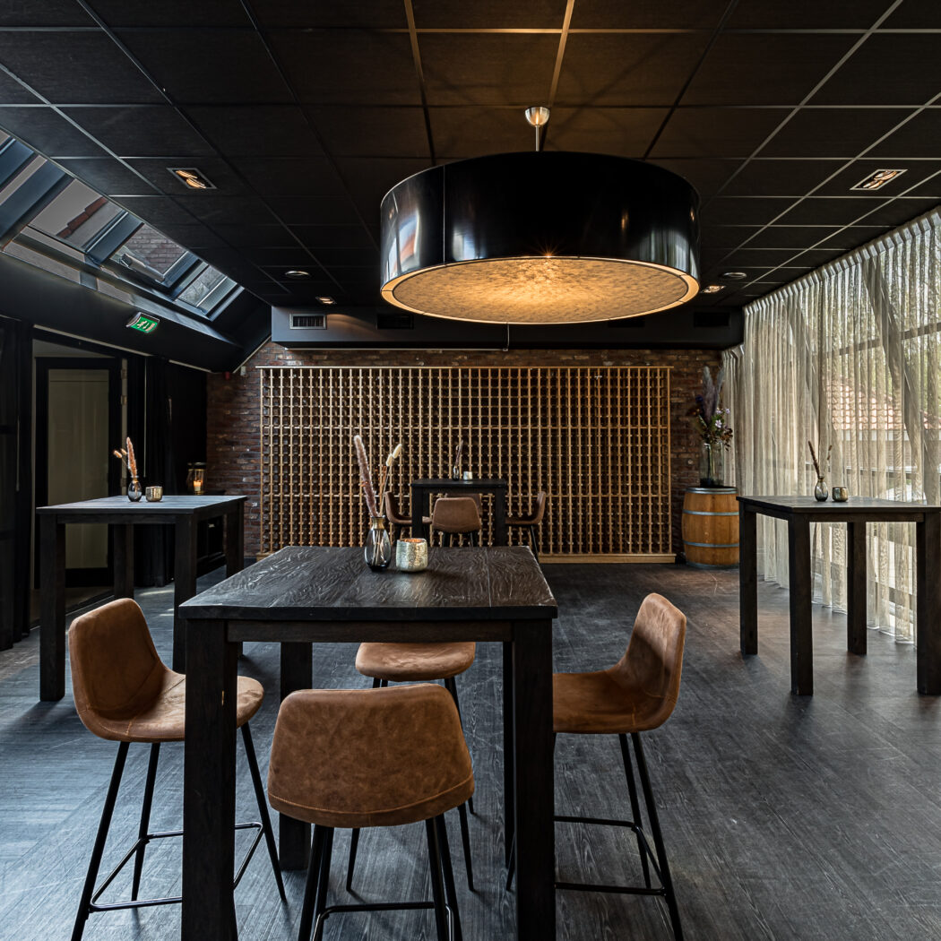 Stijlvolle evenementenlocatie met donkere tafels, bruine barkrukken, bakstenen wijnrekmuur en lichte gordijnen.