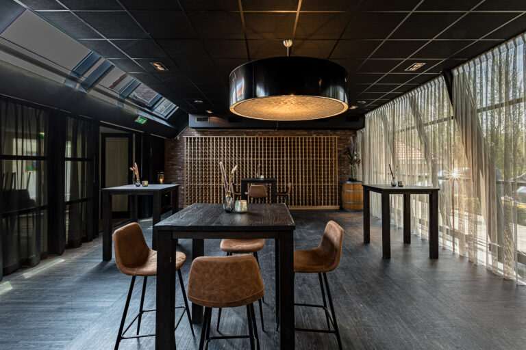 Stijlvolle evenementenlocatie met donkere tafels, bruine barkrukken, bakstenen wijnrekmuur en lichte gordijnen.