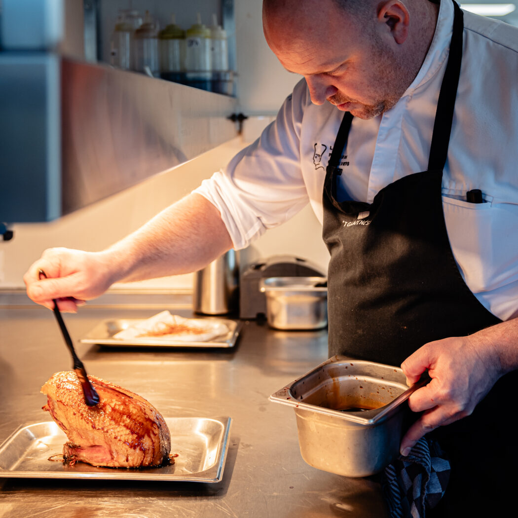 Een chef in wit uniform en zwart schort overgiet gebraden vlees met donkere saus in een professionele keuken.