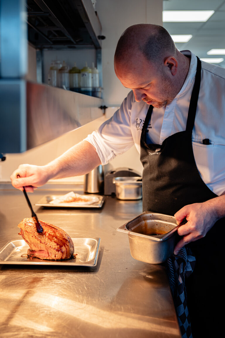 Een chef in wit uniform en zwart schort overgiet gebraden vlees met donkere saus in een professionele keuken.
