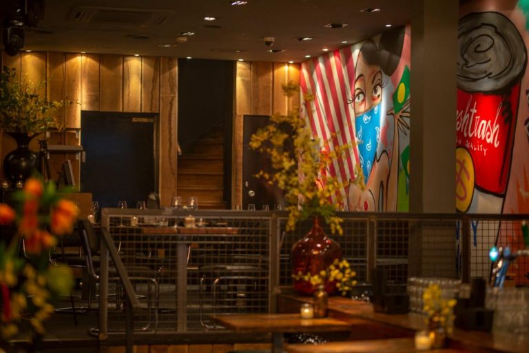 Sfeervol restaurantinterieur met houten lambrisering, tafels, bloemen en kleurrijke muurschilderingen van een vrouw en een blik met Freshtrash.