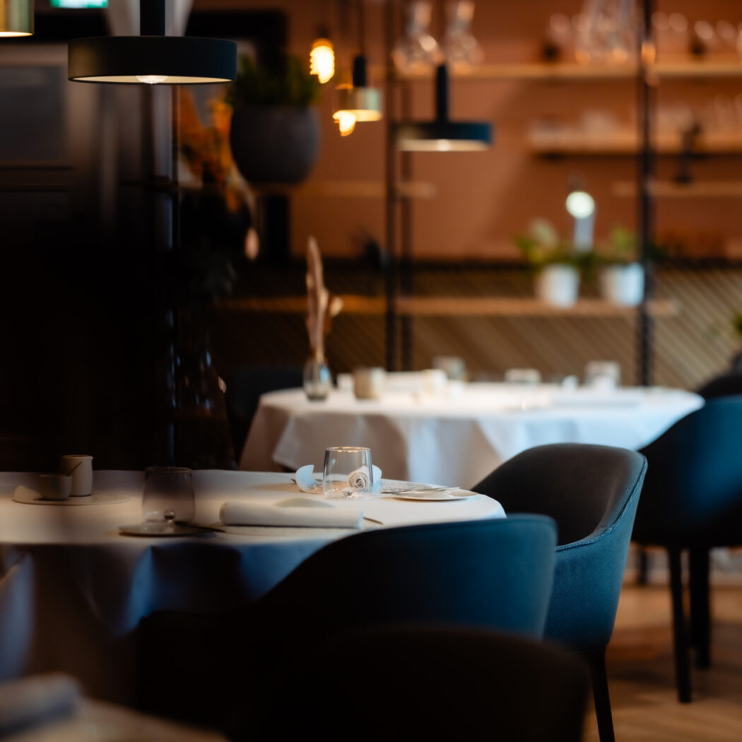 Een sfeervol restaurantinterieur met gedekte tafels, donkere stoelen en warm hanglampenlicht.