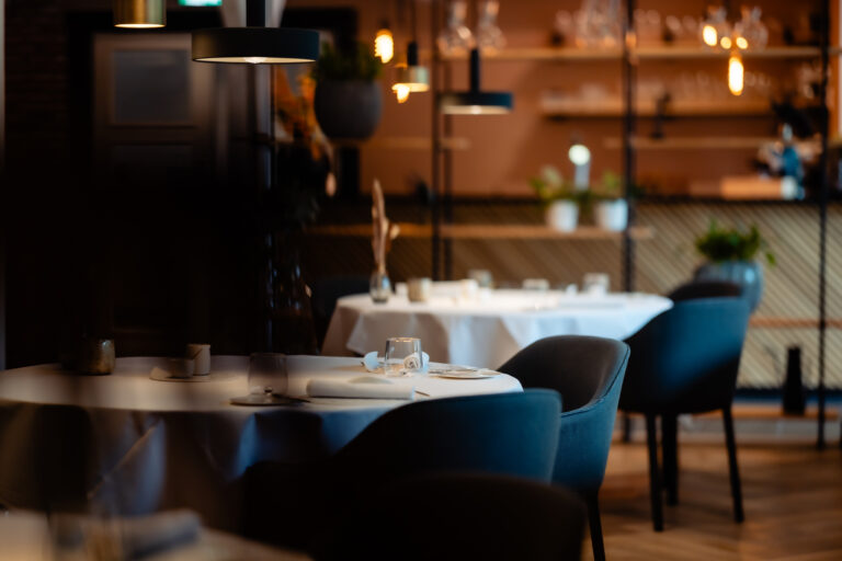 Een sfeervol restaurantinterieur met gedekte tafels, donkere stoelen en warm hanglampenlicht.