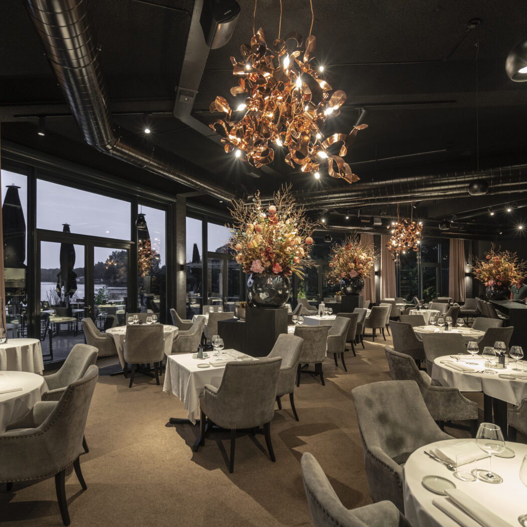 Sfeervol restaurant met gedekte tafels, koperen kroonluchters, bloemstukken en uitzicht over water in de schemer.