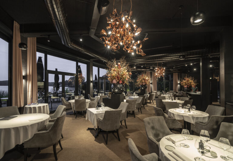 Sfeervol restaurant met gedekte tafels, koperen kroonluchters, bloemstukken en uitzicht over water in de schemer.