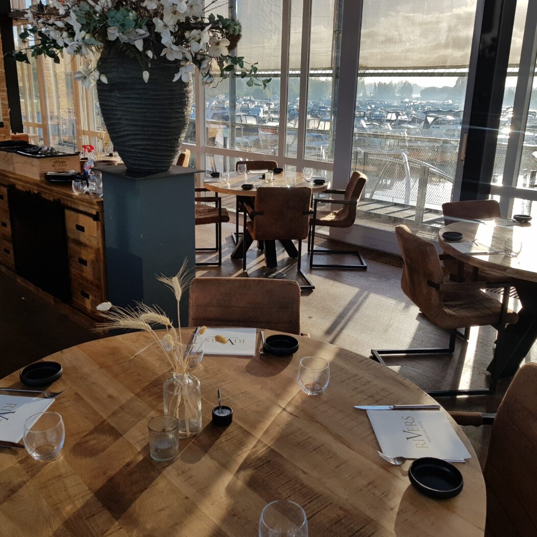Modern restaurantinterieur met houten tafels, RIVERS menus en zonnig uitzicht over een jachthaven.