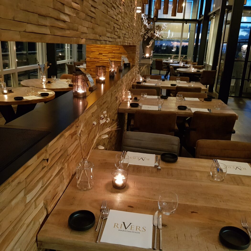 Sfeervol restaurant met houten tafels, kaarslicht, stenen muur en servetten met RIVERS.