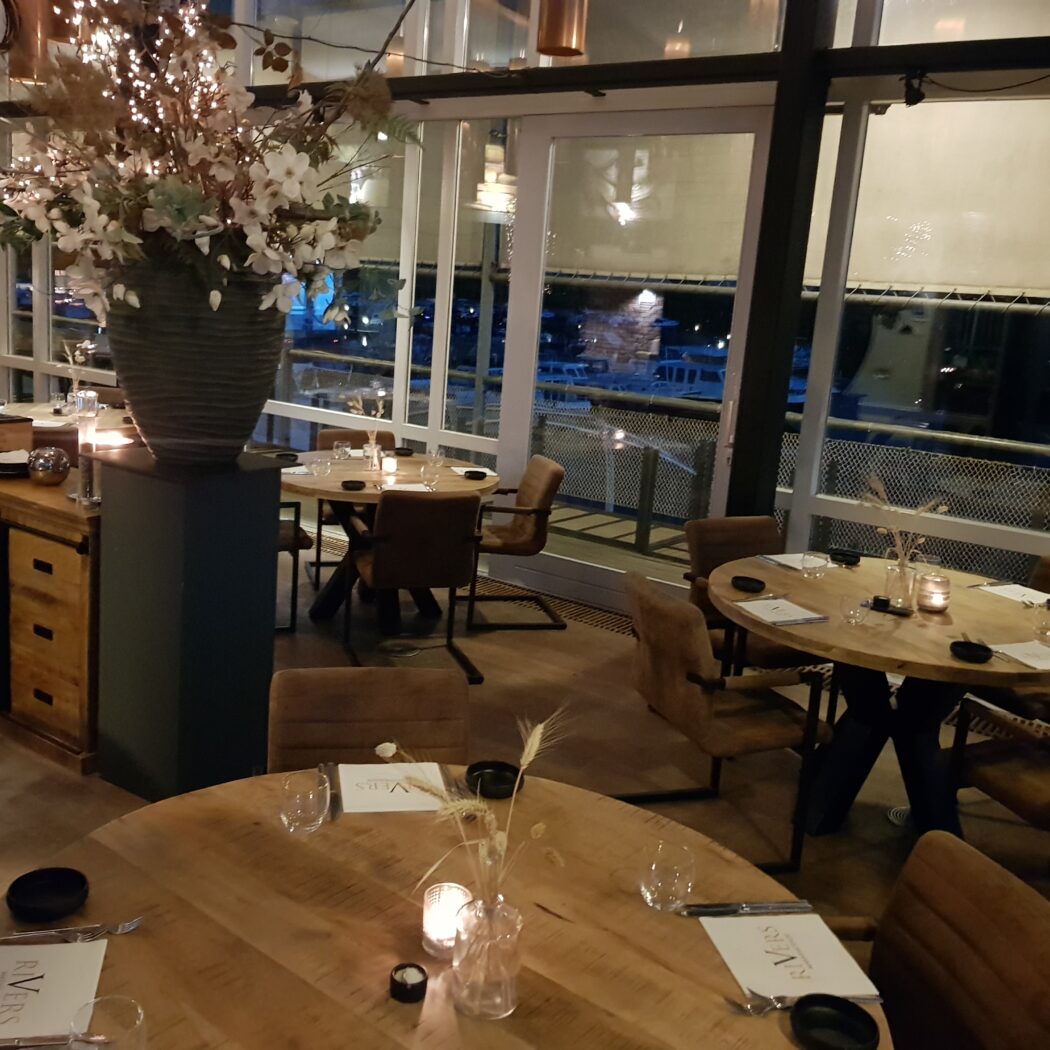 Sfeervol restaurant bij avond met houten tafels, RIVERS menus, grote ramen en verlichte bloemenvase.
