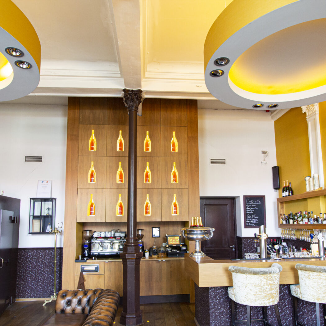 Sfeervolle bar met gele muren, houten toog, verlichte flessenwand en ovale plafondlampen.