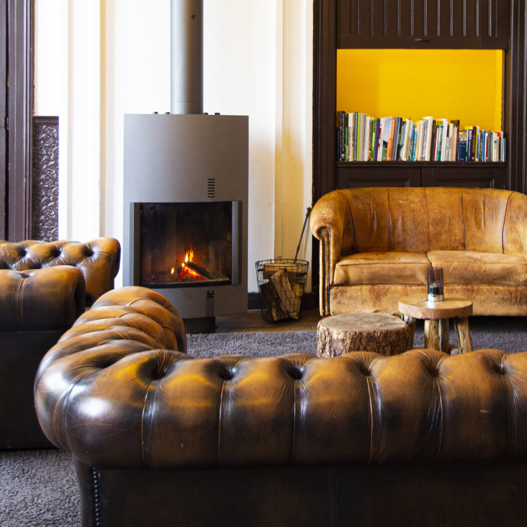 Sfeervolle lounge met brandende haard, bruine Chesterfield banken en een gele boekenkast.