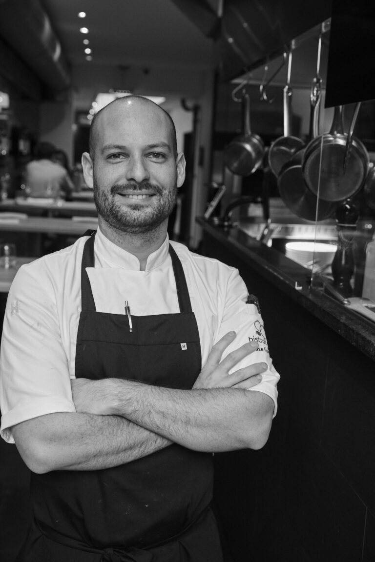 Lachende kok met baard en koksuniform, armen gekruist, in een restaurantkeuken.