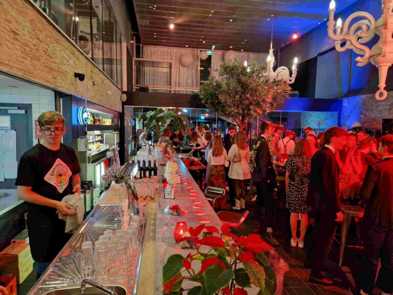 Jonge barman in bril en Brouwerij t IJ T-shirt veegt lange bar schoon te midden van feestende gasten.