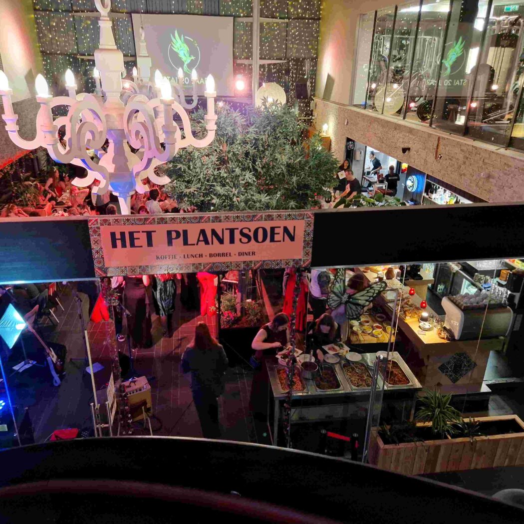Drukbezocht evenement in Het Plantsoen met witte kroonluchter, veel groen, een band en mensen bij de bar en buffet.
