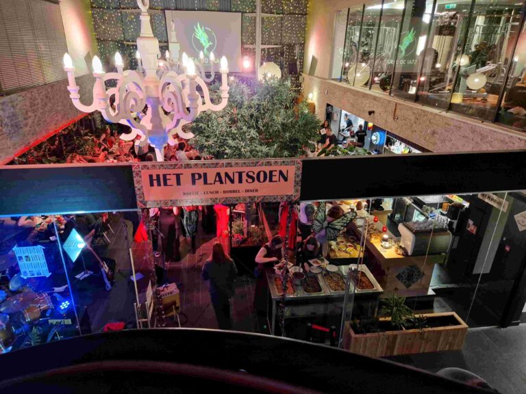 Drukbezocht evenement in Het Plantsoen met witte kroonluchter, veel groen, een band en mensen bij de bar en buffet.