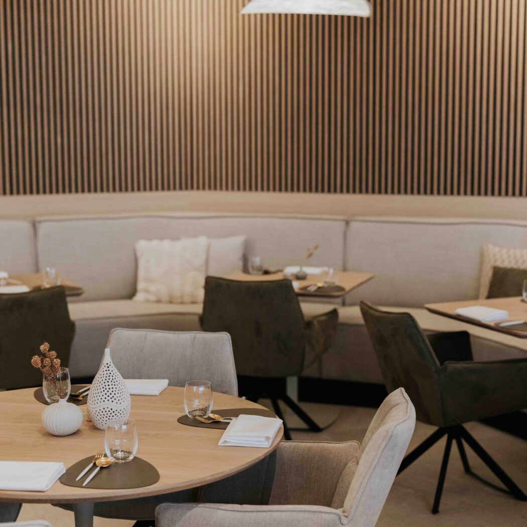 Modern restaurant interieur met ronde houten tafels, beige bank en stoelen, lattenwand en witte lampen.