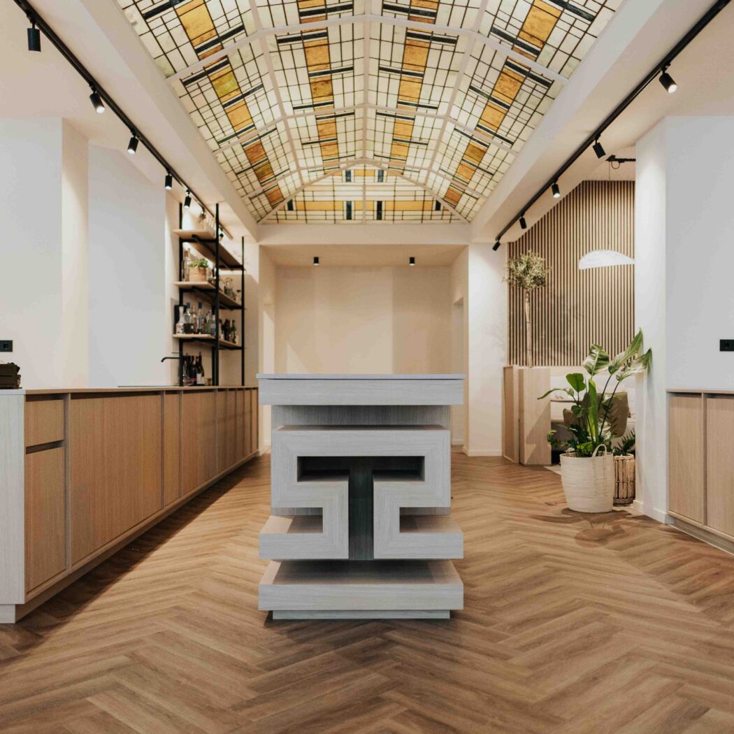 Modern interieur met visgraatvloer, lange houten balie, centraal geometrisch eiland en kleurrijk glazen plafond.