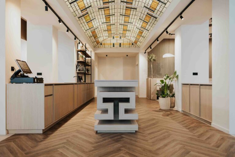 Modern interieur met visgraatvloer, lange houten balie, centraal geometrisch eiland en kleurrijk glazen plafond.