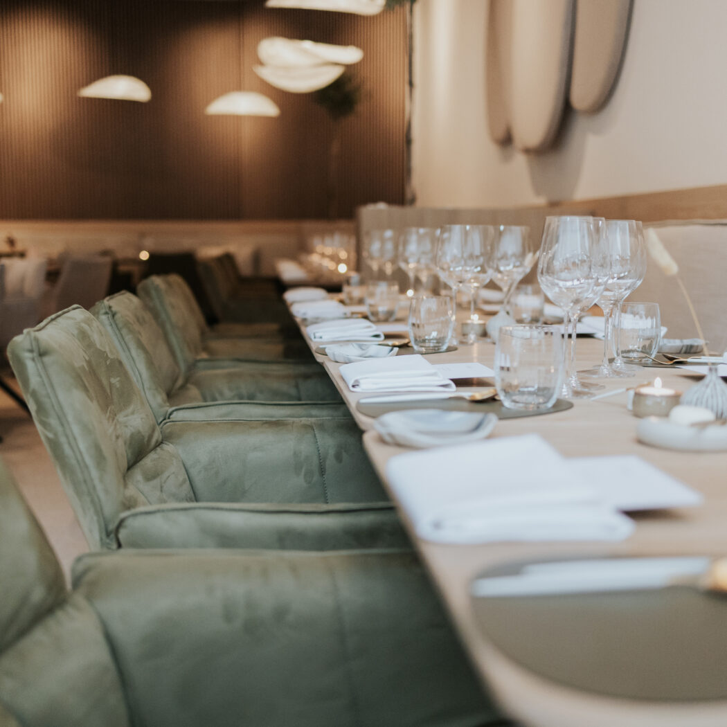 Lange eettafel in een elegant restaurant, gedekt met glazen, gouden bestek, witte servetten en groene stoelen.