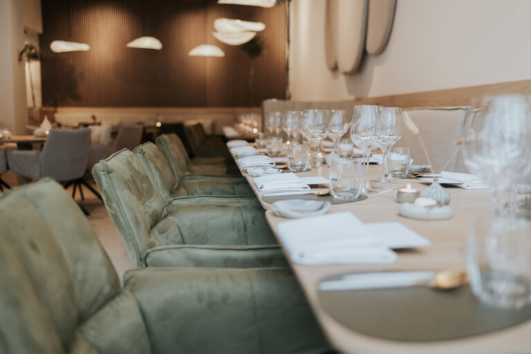 Lange eettafel in een elegant restaurant, gedekt met glazen, gouden bestek, witte servetten en groene stoelen.