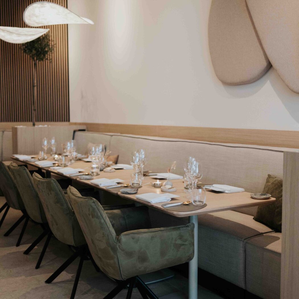 Modern restaurantinterieur met lange, gedekte tafel, olijfgroene stoelen en beige zitbank.