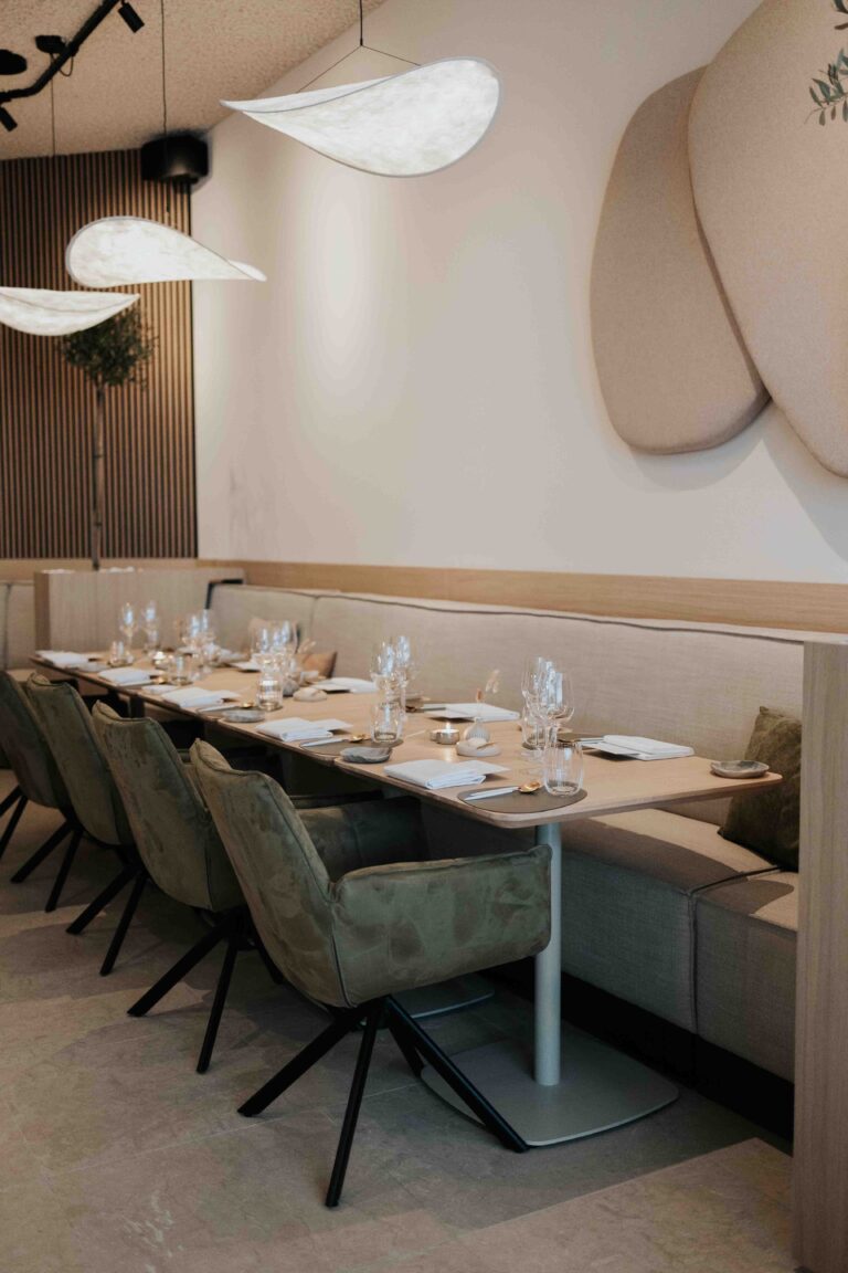 Modern restaurantinterieur met lange, gedekte tafel, olijfgroene stoelen en beige zitbank.