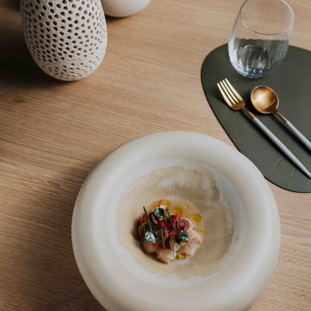 Culinair gerecht met rauwe vis en garnering in een lichtbeige kom op een houten tafel, met goudkleurig bestek.