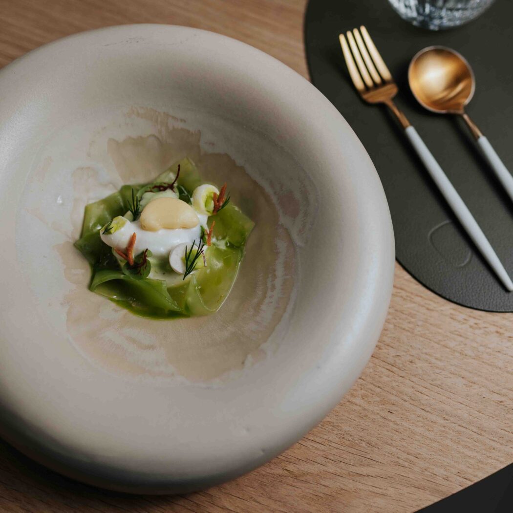 Fijnproeversgerecht met groene linten, witte crème en gele topping in een beige kom, naast goudkleurig bestek op houten tafel.