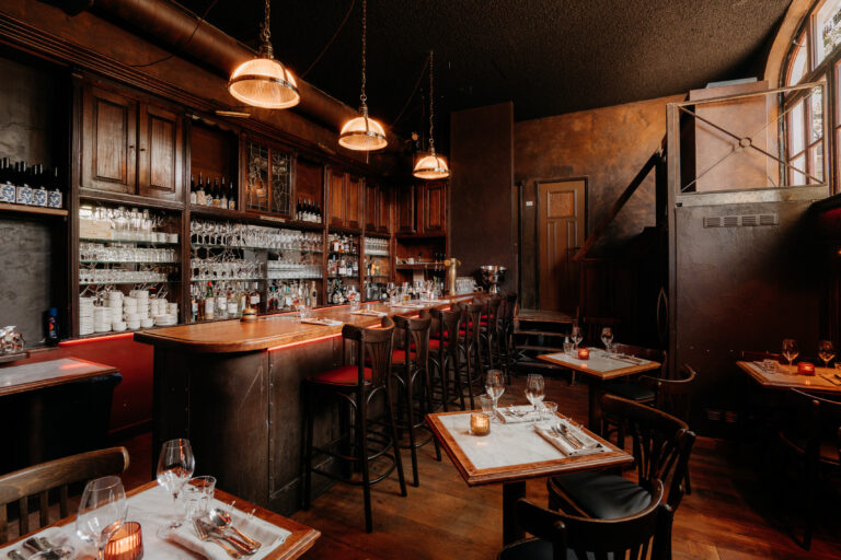 Sfeervol restaurant interieur met een lange donkerhouten bar, barkrukken, en gedekte tafels met kaarsen.