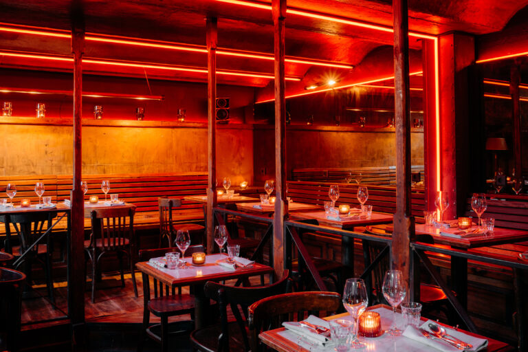 Donker restaurantinterieur met rode neon accenten, houten meubels en gedekte tafels met wijnglazen en theelichtjes.