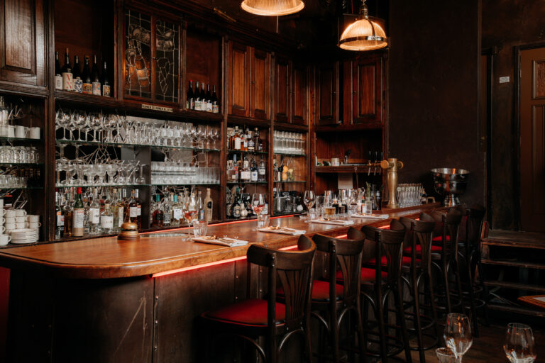 Sfeervolle klassieke bar met houten toonbank, rode accentverlichting, barkrukken en donkere kasten vol drankflessen en glazen.