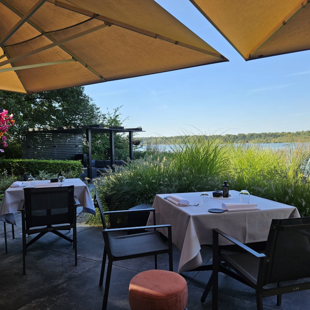 Buitenterras met beige parasols, gedekte tafels, kleurrijke bloemen en weids uitzicht op water en groene oever.