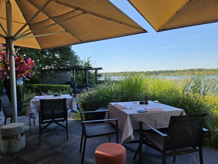 Buitenterras met beige parasols, gedekte tafels, kleurrijke bloemen en weids uitzicht op water en groene oever.