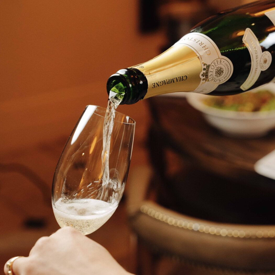 Een hand schenkt Christoffe Champagne uit een groene fles met goud etiket in een flute glas, waarbij bubbels ontstaan.