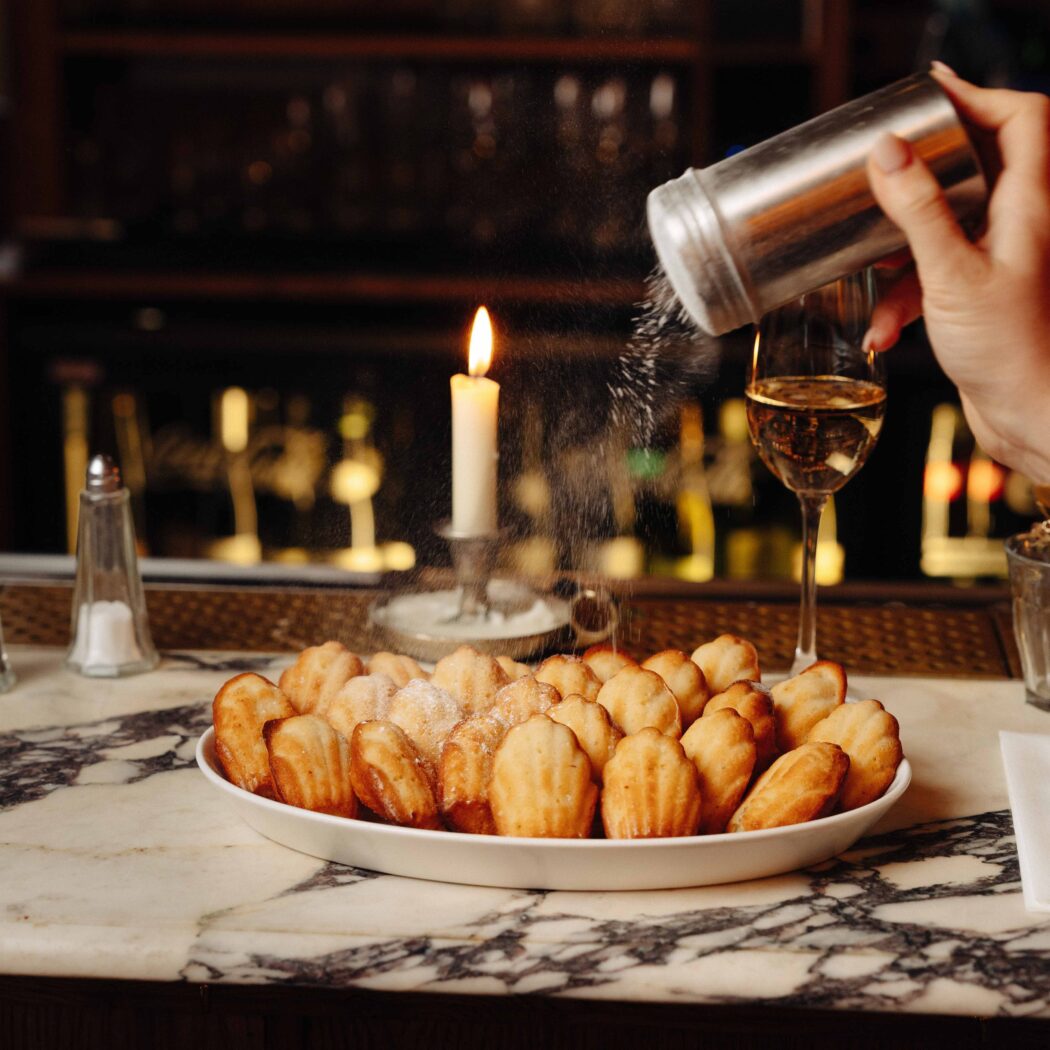 Een hand strooit poedersuiker over een schaal madeleines op een marmeren bar, met een brandende kaars en glas wijn.