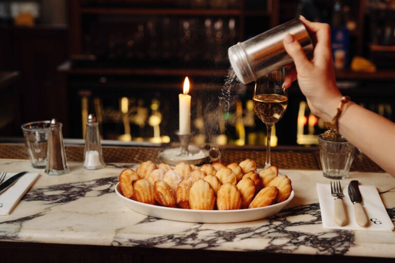 Een hand strooit poedersuiker over een schaal madeleines op een marmeren bar, met een brandende kaars en glas wijn.