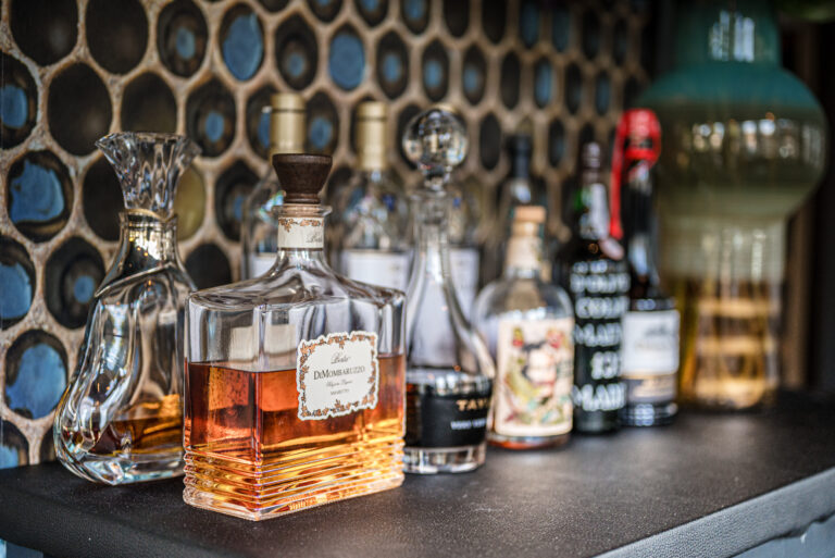 Vierkante fles Porta DMOMBARUZZO Amaretto en een glazen karaf op een bar voor een decoratieve tegelwand.
