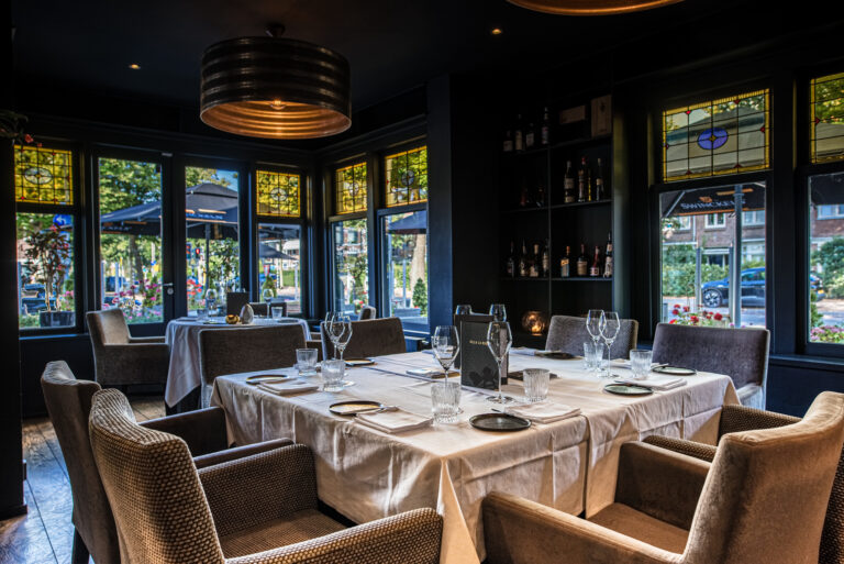 Gezellig restaurantinterieur met gedekte tafels, grote ramen met gebrandschilderd glas en menukaart “VILLA LA RO...”