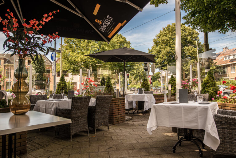 Gezellig restaurantterras met gedekte tafels, rieten stoelen, bloemen en zwarte parasols, waaronder een Swinckels parasol.