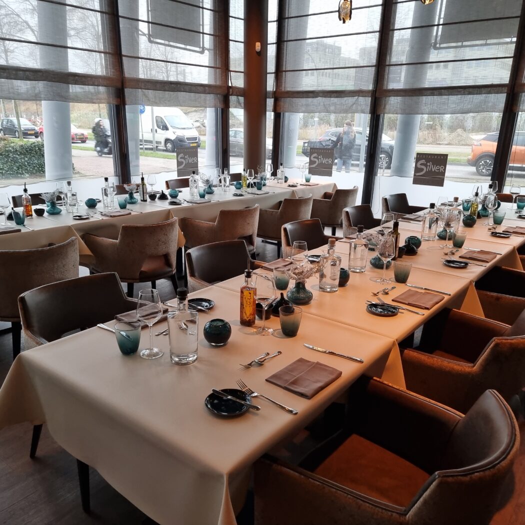 Interieur van Restaurant Silver met meerdere lange, gedekte tafels, bruine stoelen en grote ramen met uitzicht op de weg.