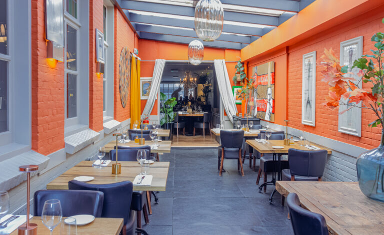 Lichte restaurant serre met oranje bakstenen muren, houten tafels, donkere stoelen en grote glazen hanglampen.