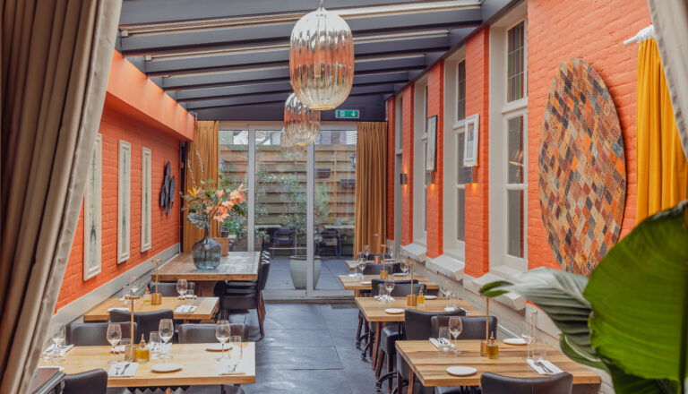 Sfeervol restaurantinterieur met oranje bakstenen muren, gedekte houten tafels, designlampen en uitzicht op een binnentuin.