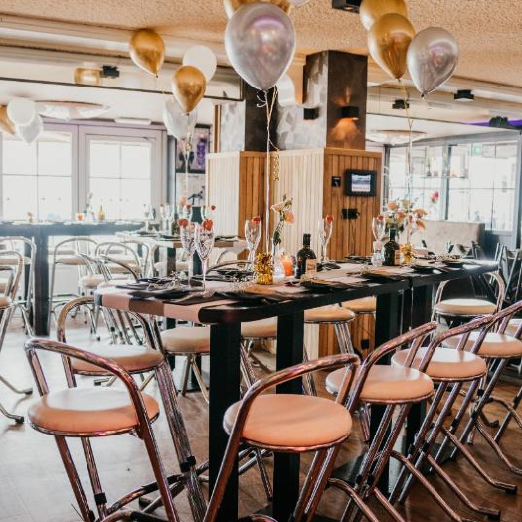 Feestelijk gedekte hoge tafels met crème krukken en gouden, zilveren en witte ballonnen in een evenementenruimte.